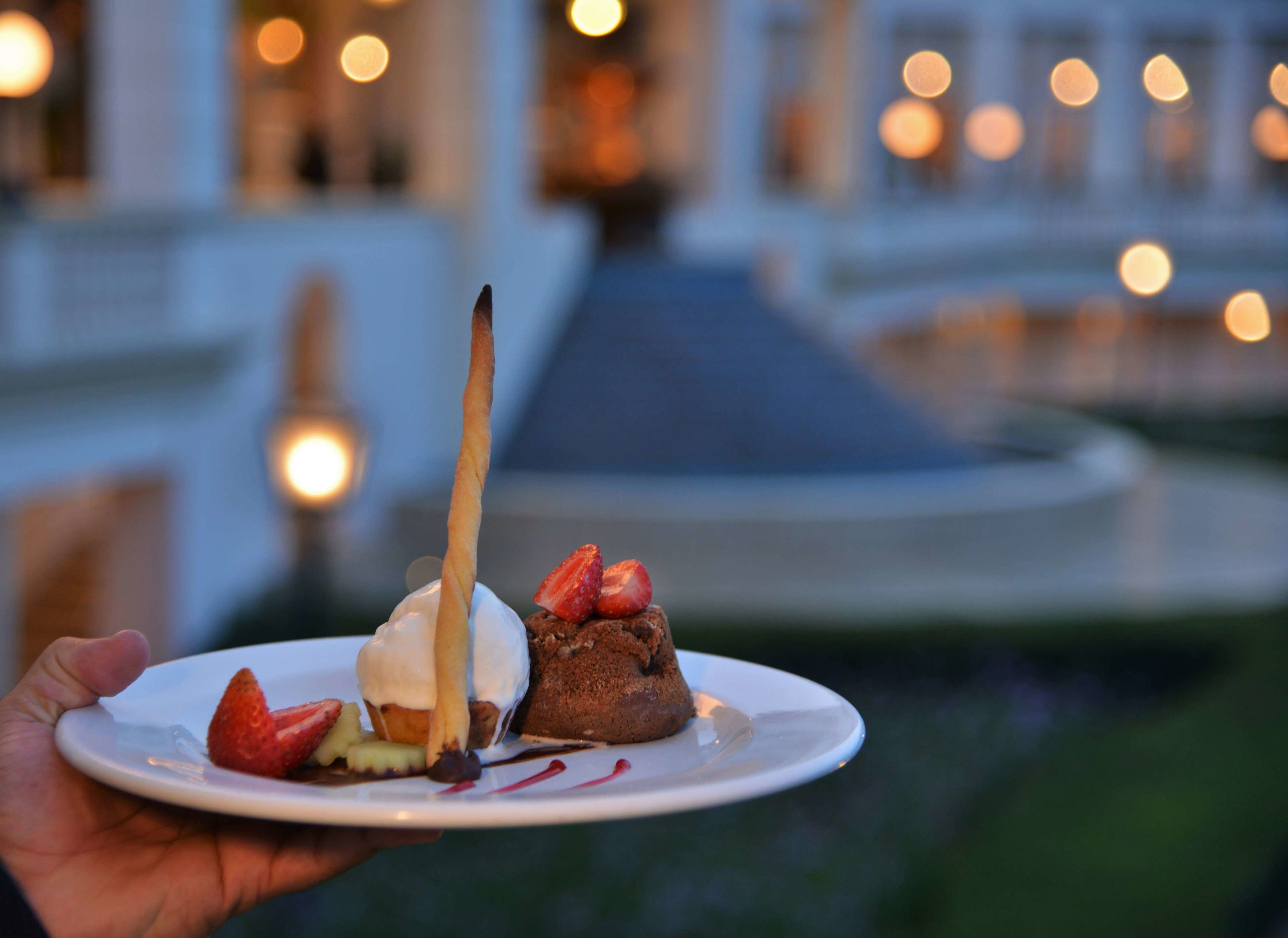 A dessert plate with a chocolate cake, strawberries, and a chocolate sauce, held in front of a building with lights in the background.