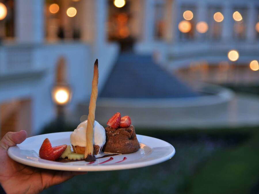 A dessert plate with a chocolate cake, strawberries, and a chocolate sauce, held in front of a building with lights in the background.
