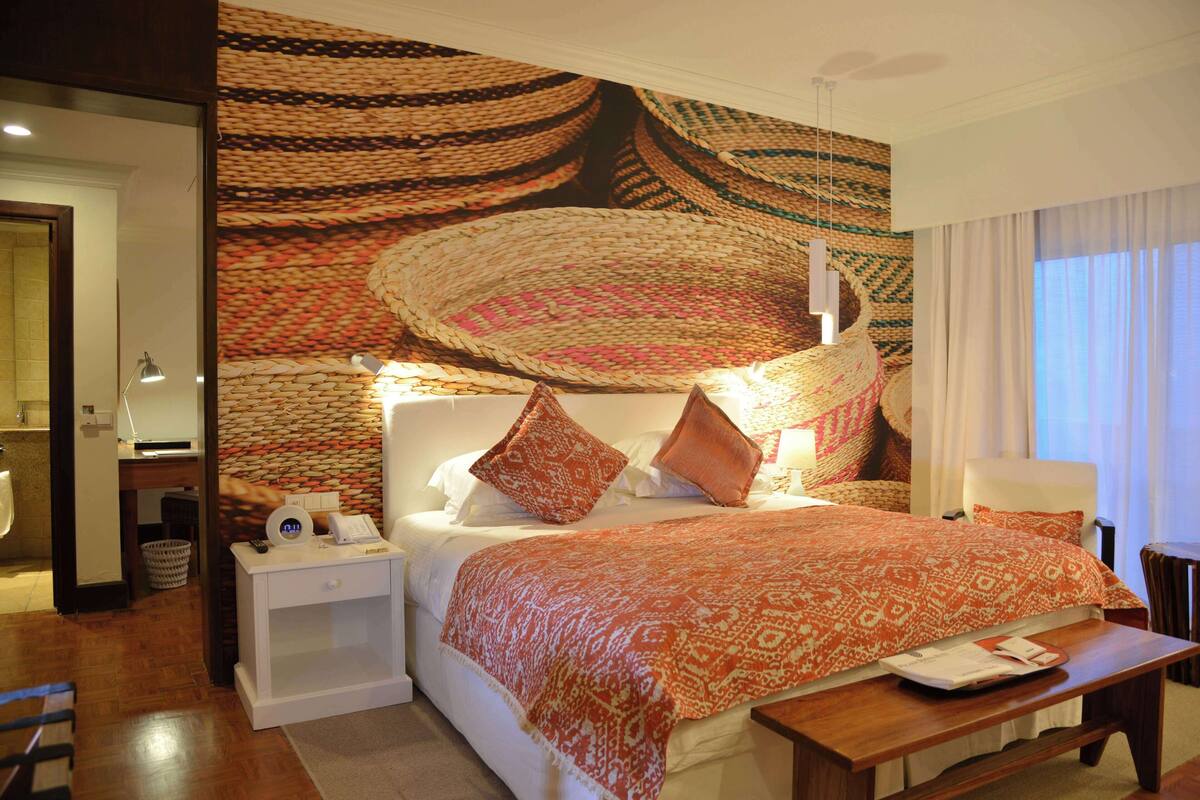 Ein Hotelzimmer mit einer großen, gewebten Wandtextil, einem Bett mit gemusterten Bettbezügen, einem Nachttisch mit einer Lampe und einem Holzbeistelltisch.