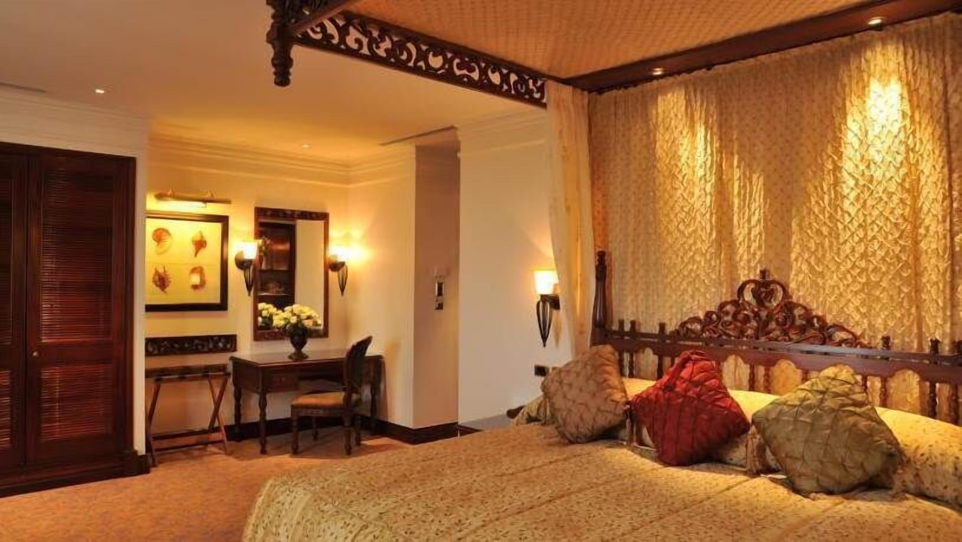 A bedroom with a large bed, a desk with a chair, a mirror, and wall sconces.
