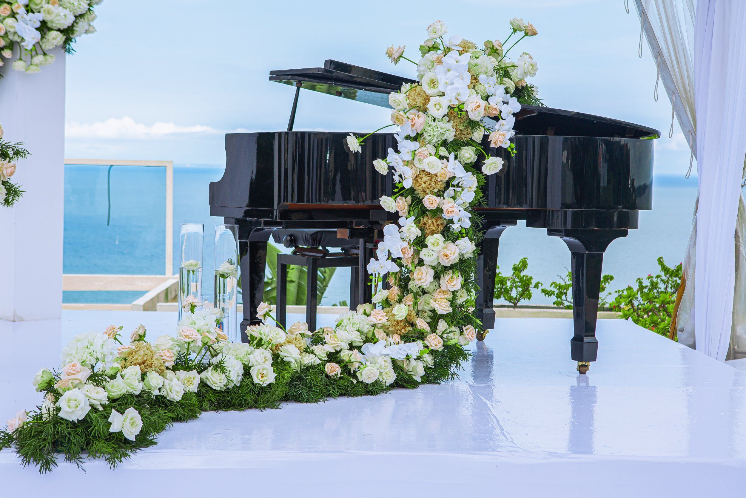 A grand piano on a stage with a floral arrangement and a view of the sea.