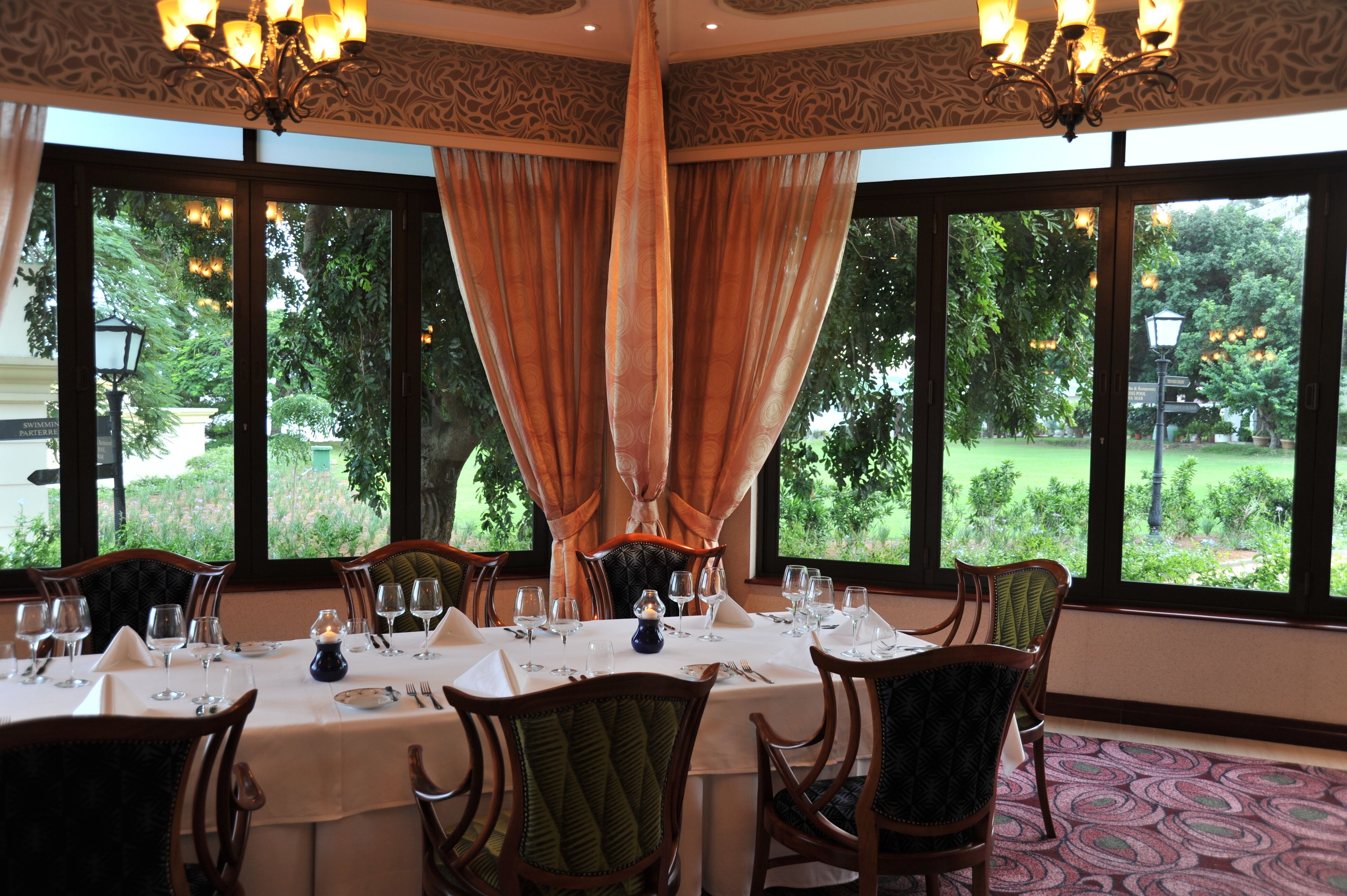 A dining area with a table set for six, large windows with curtains, and a chandelier.