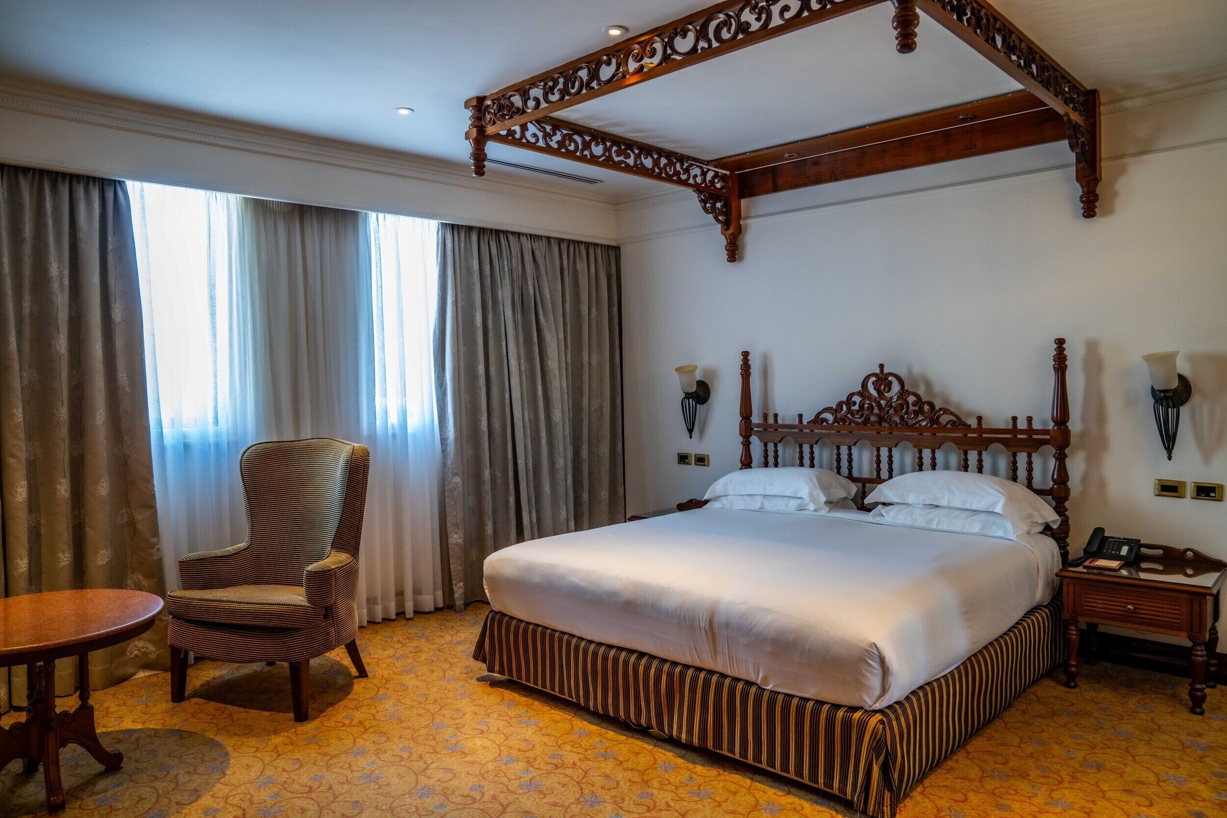 A hotel room with a large bed, a chair, a table, and a canopy bed frame.