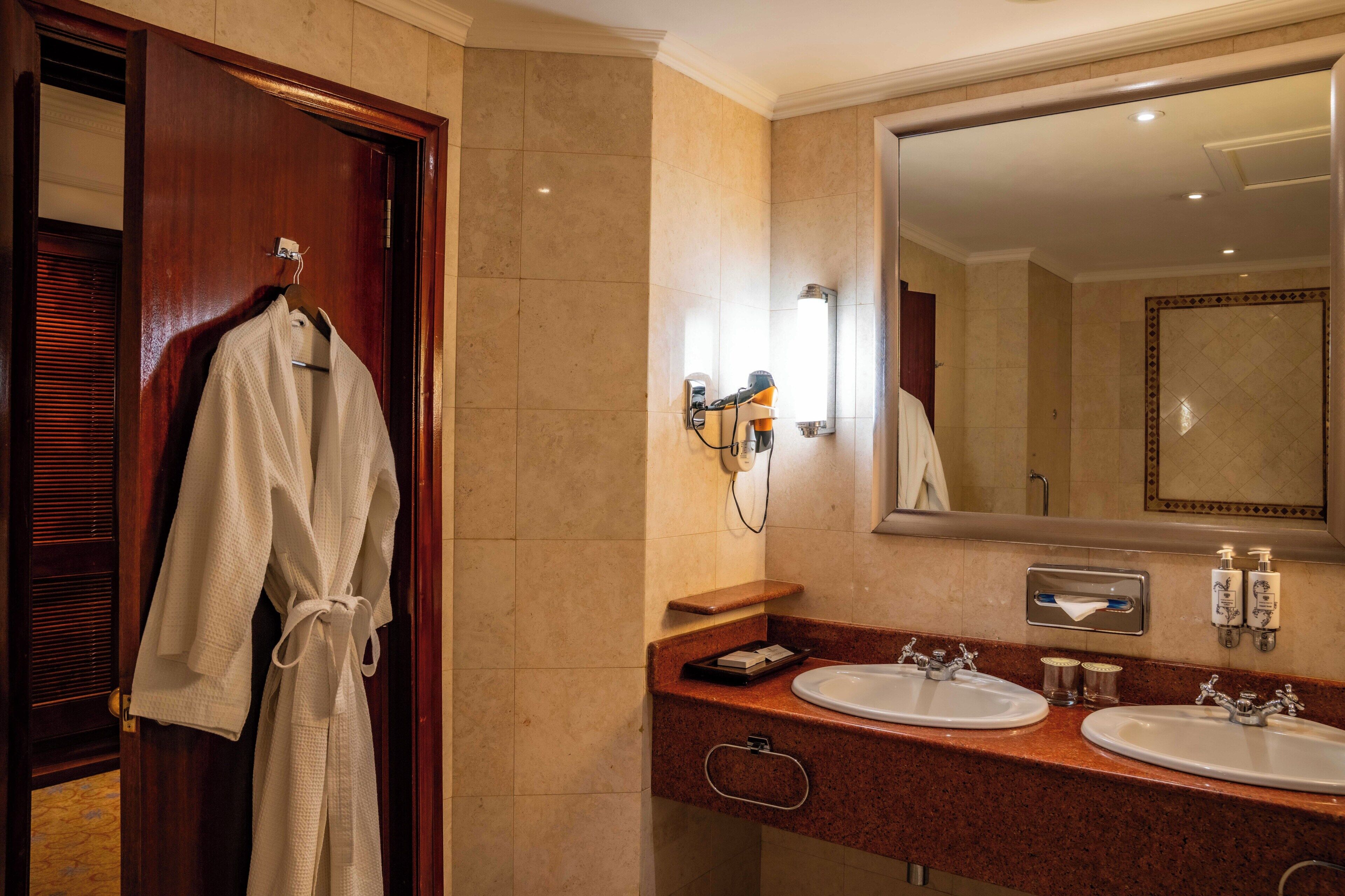 A hotel bathroom with a wooden door, a robe hanging on a rack, a large mirror, two sinks, and toiletries.