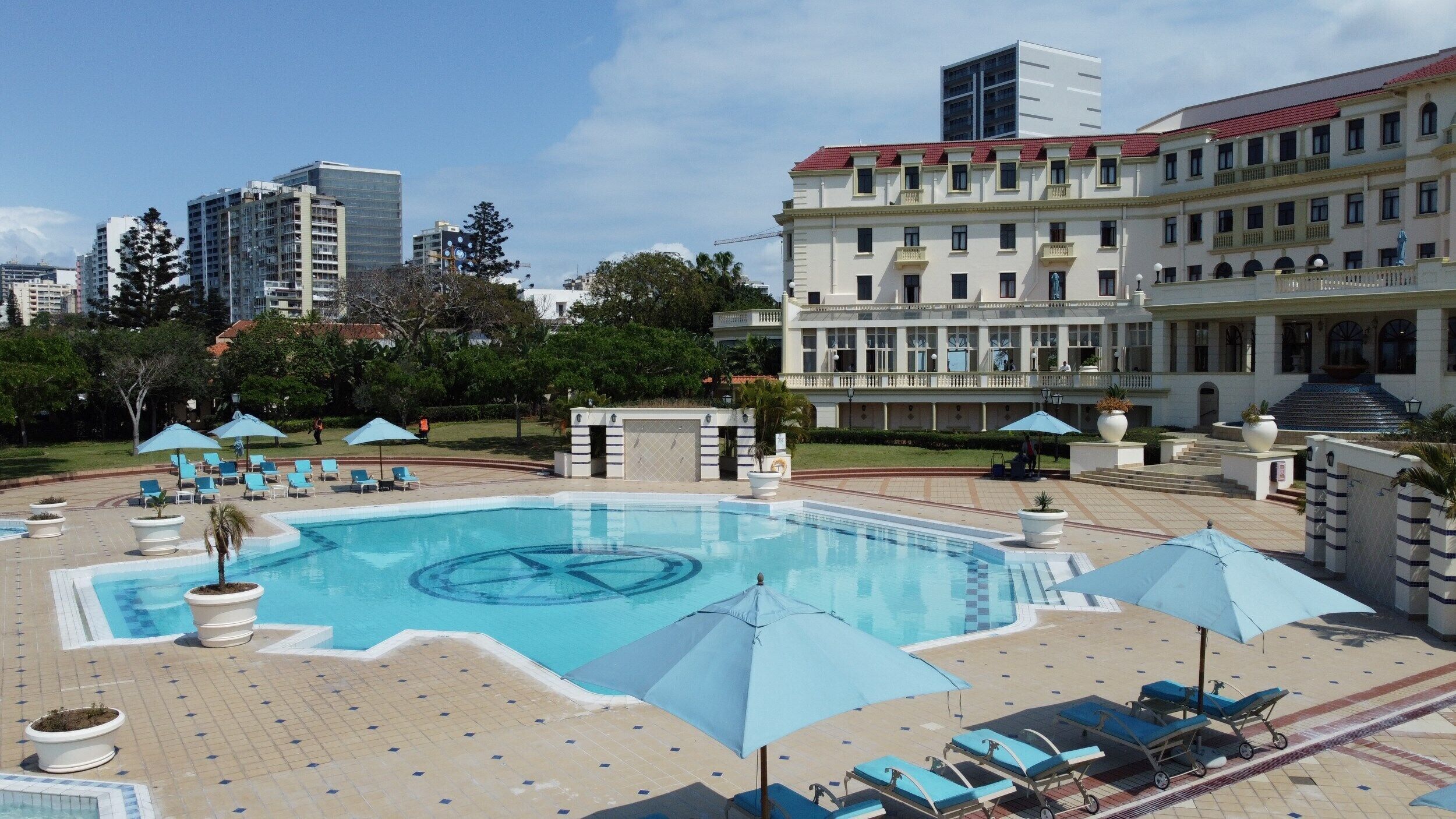 A swimming pool with lounge chairs and umbrellas, surrounded by a landscaped area and a large building with multiple floors.