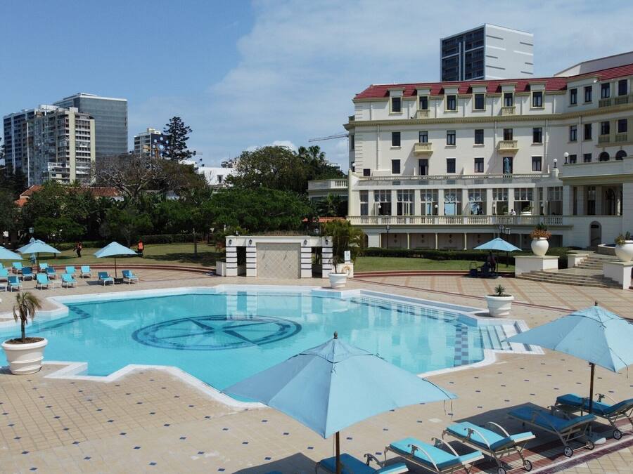 A swimming pool with lounge chairs and umbrellas, surrounded by a landscaped area and a large building with multiple floors.