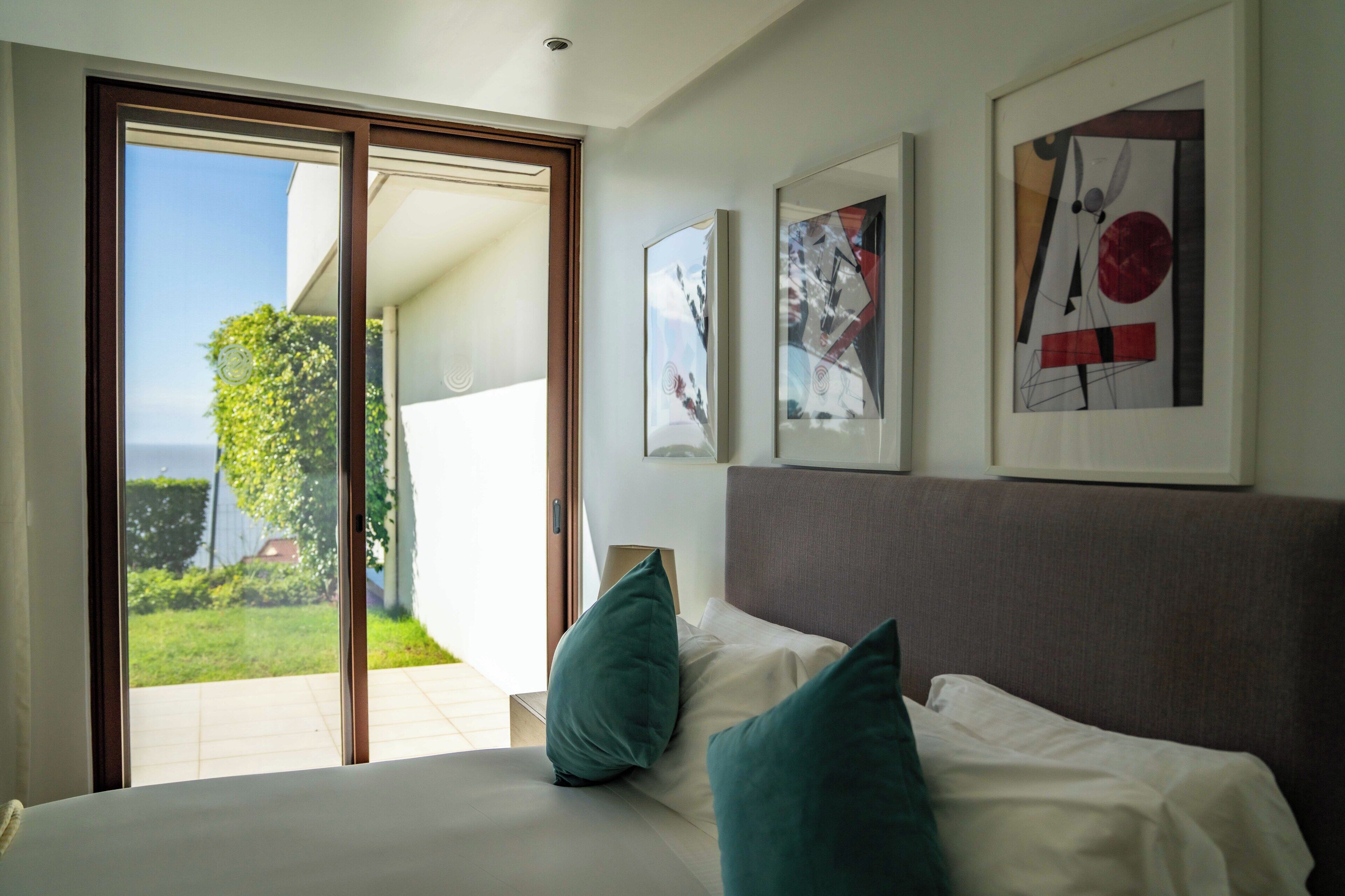 Un dormitorio con cama, almohadas, una mesita de noche y un ventanal con vista a un jardín y un edificio.