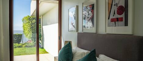 Un dormitorio con cama, almohadas, una mesita de noche y un ventanal con vista a un jardín y un edificio.