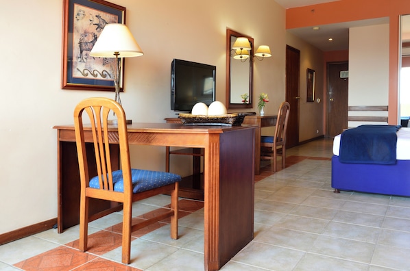 A hotel room with a wooden desk, a chair, a television, a bed, and a bathroom visible in the background.