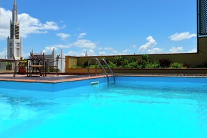 Una piscina con una escalera metálica, una silla y una mesa. Al fondo, se observa un edificio alto con una torre de reloj.