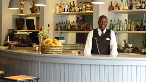 A bar area with a counter, shelves of bottles, and a seated area with a chair and table. - Pestana Rovuma (Maputo)