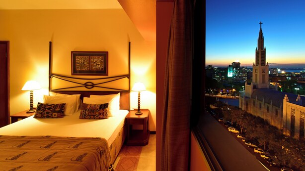 A hotel room with a bed, two bedside lamps, a nightstand, and a view of a cityscape with a church tower. - Pestana Rovuma (Maputo)