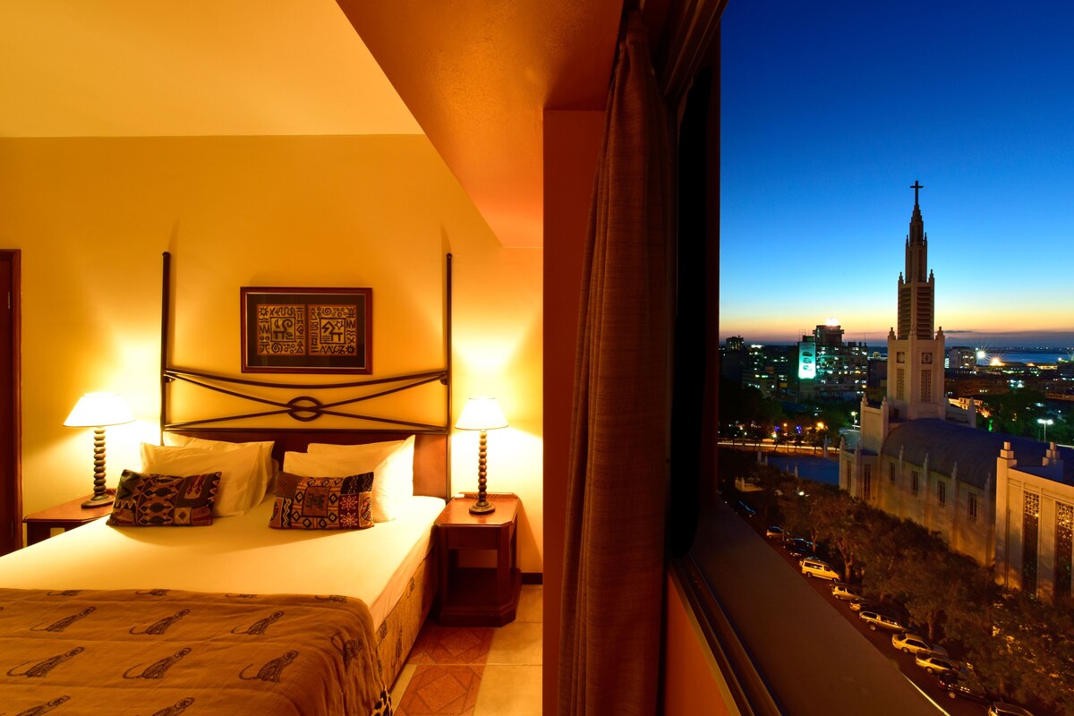 A hotel room with a bed, two bedside lamps, patterned pillows, and a view of a cityscape with a prominent church tower.