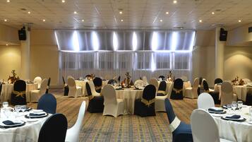 Un salón de banquetes con mesas dispuestas para un evento formal, con alfombra estampada y un gran ventanal con cortinas traslúcidas.