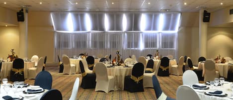 Un salón de banquetes con mesas dispuestas para un evento formal, con alfombra estampada y un gran ventanal con cortinas traslúcidas.