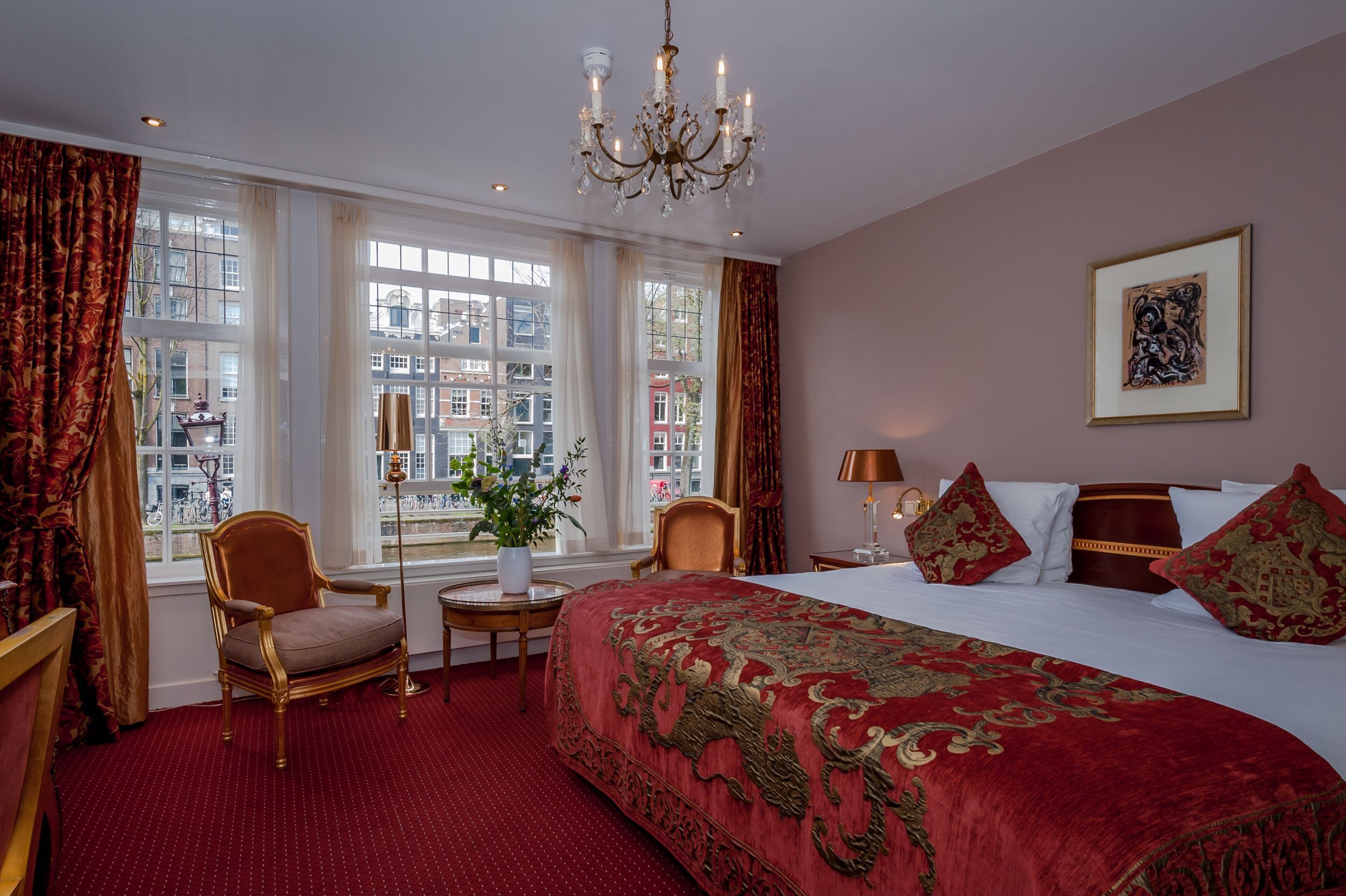 Una habitación de hotel con una cama grande, un candelabro, vistas de la ciudad a través de amplios ventanales y un suelo cubierto de alfombra roja.