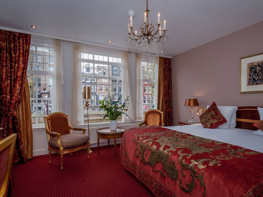 Una habitación de hotel con una cama grande, un candelabro, vistas de la ciudad a través de amplios ventanales y un suelo cubierto de alfombra roja.