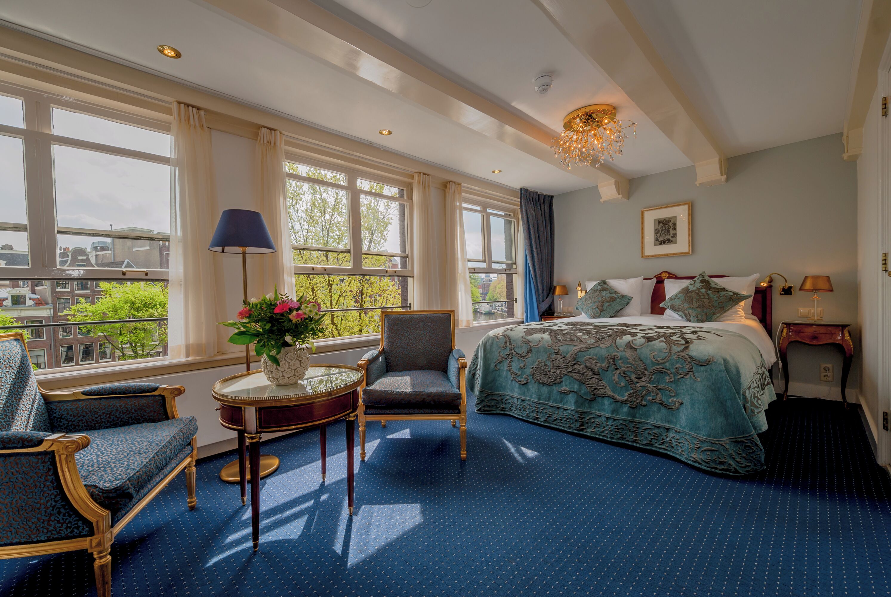 Quarto de hotel com uma cama grande, área de estar com uma poltrona e uma mesa pequena, e vista da cidade através de janelas amplas.