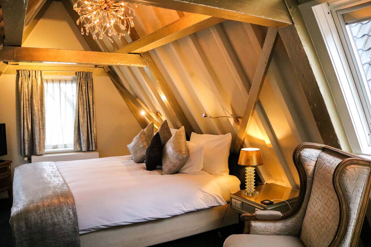 Quarto de hotel com teto alto, cama com roupa de cama branca, mesa de cabeceira com abajur e uma cadeira.