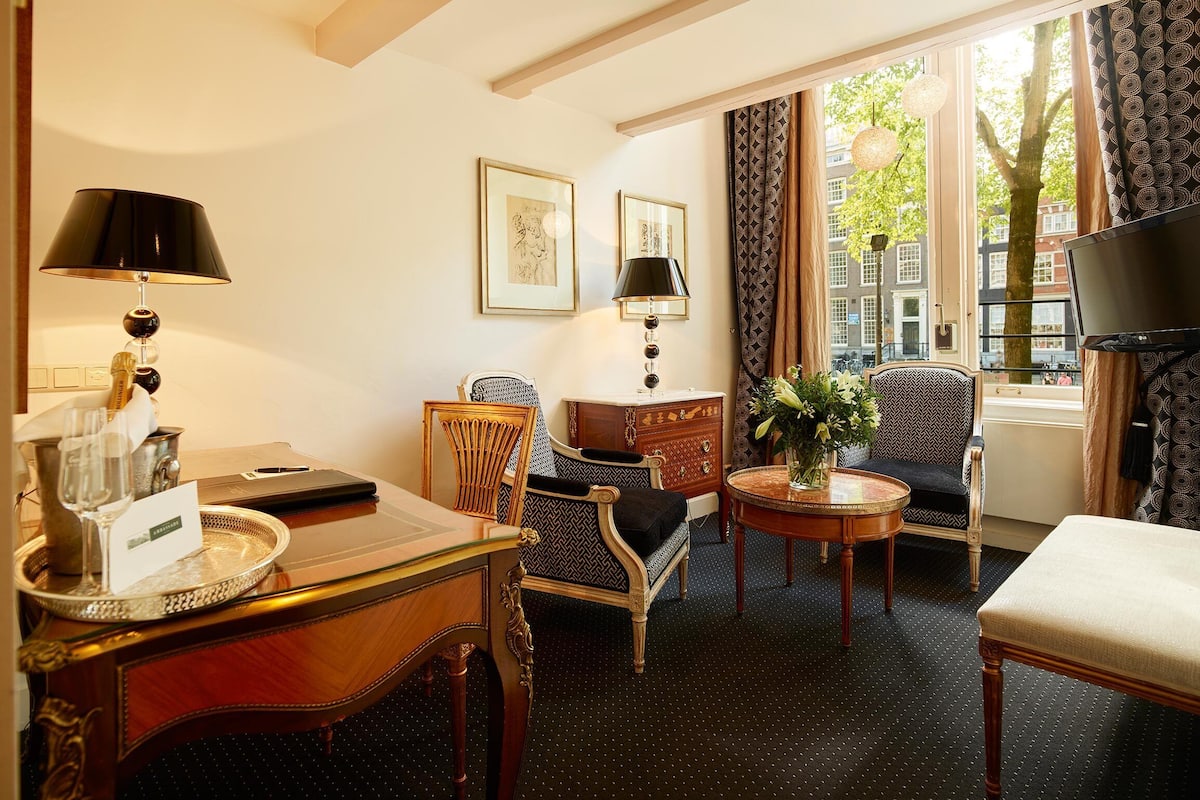 Quarto de hotel com escrivaninha de madeira, duas cadeiras, um sofá, uma mesa pequena com um vaso de flores, uma televisão e uma janela com cortinas.