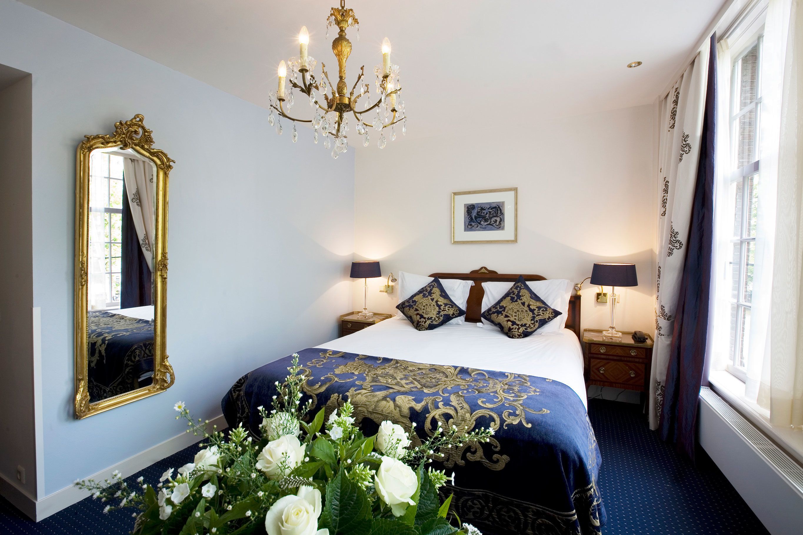 Habitación de hotel con una cama grande, un candelabro, un espejo, un cuadro en la pared y una ventana con cortinas.