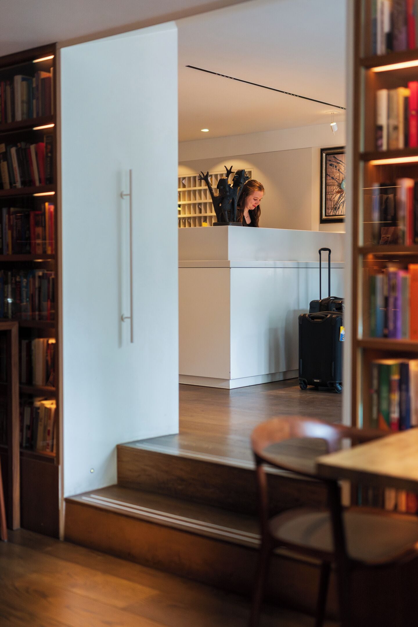 A hotel room with a wooden floor, a dining table with chairs, a bookshelf, and a suitcase on the floor.
