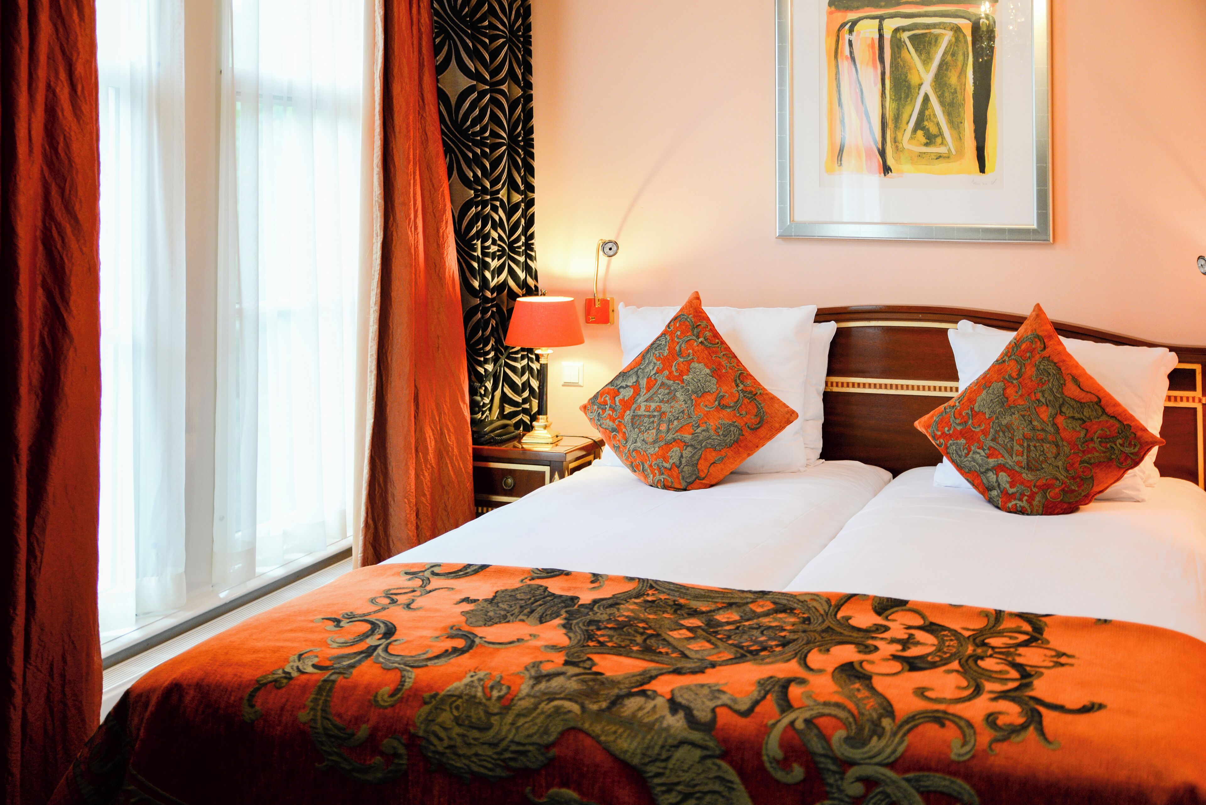 A hotel room with a large bed, two pillows, a nightstand, a lamp, and a window with curtains.