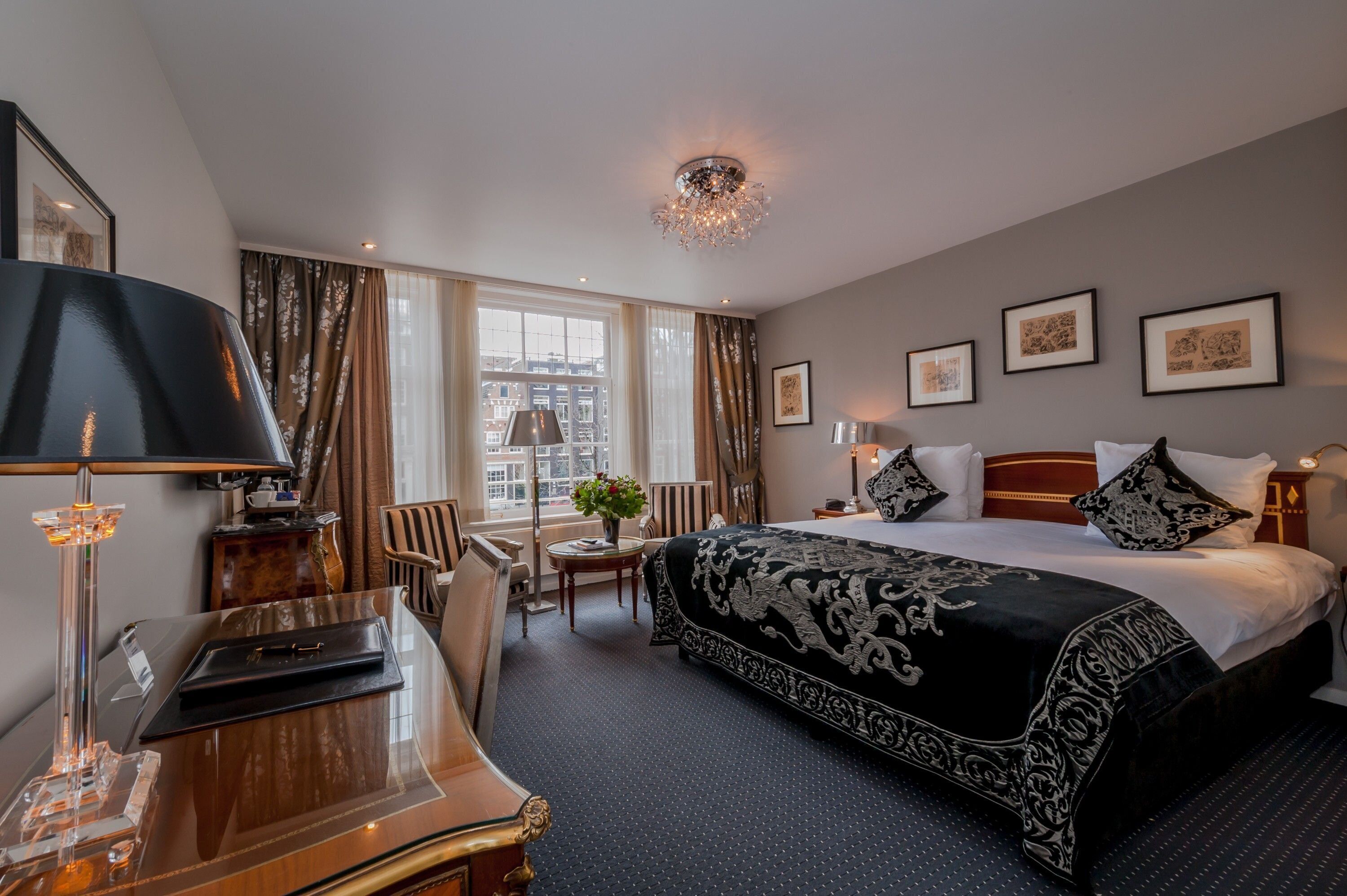 A hotel room with a large bed, a desk with a lamp, a chair, a small table with a vase, and a view of the city through large windows.
