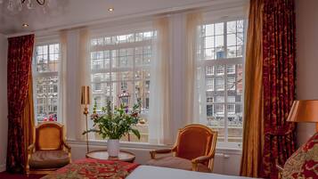 Une chambre d’hôtel avec un grand lit, deux fauteuils, un vase de fleurs et une vue sur la ville grâce à de grandes fenêtres.