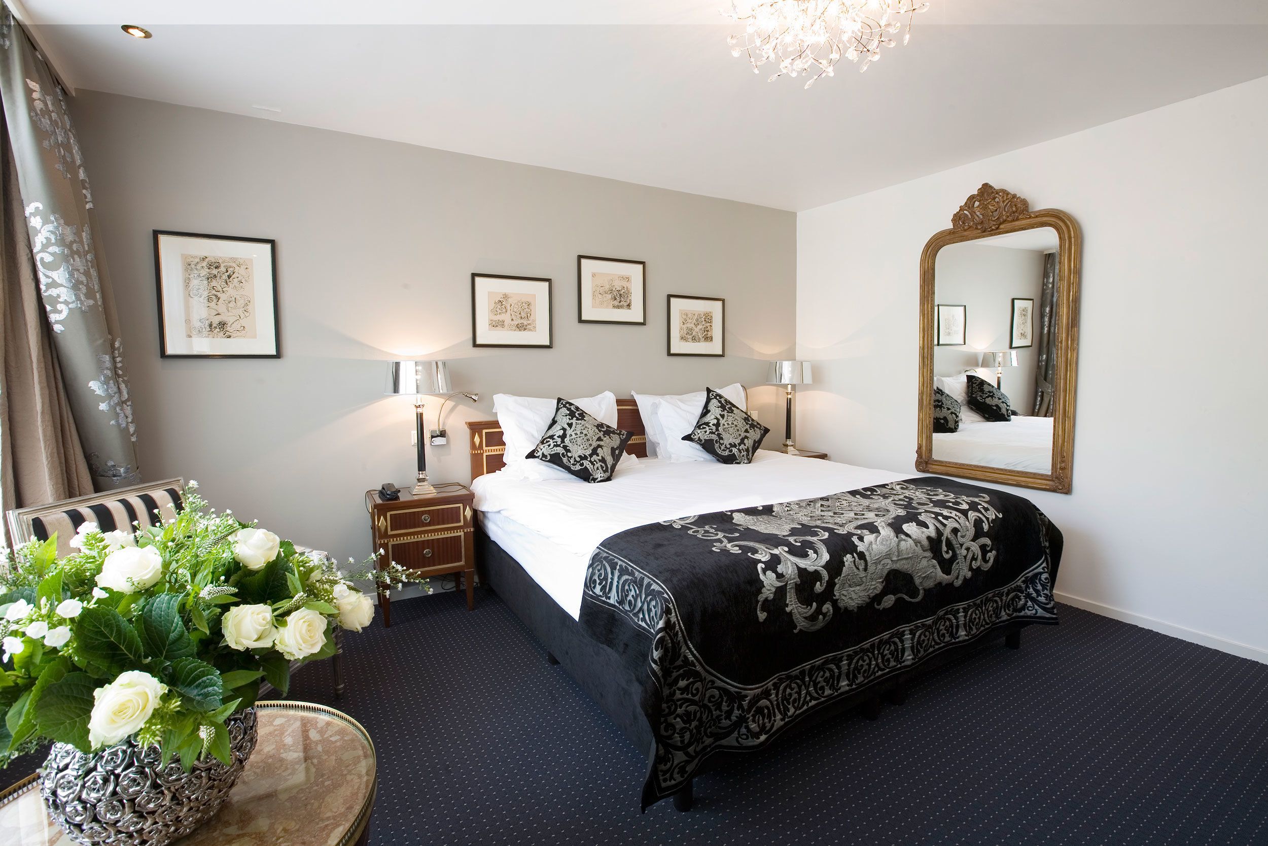 Une chambre d’hôtel avec un grand lit, un miroir, des œuvres d’art encadrées accrochées au mur et un vase de fleurs blanches posé sur une table.