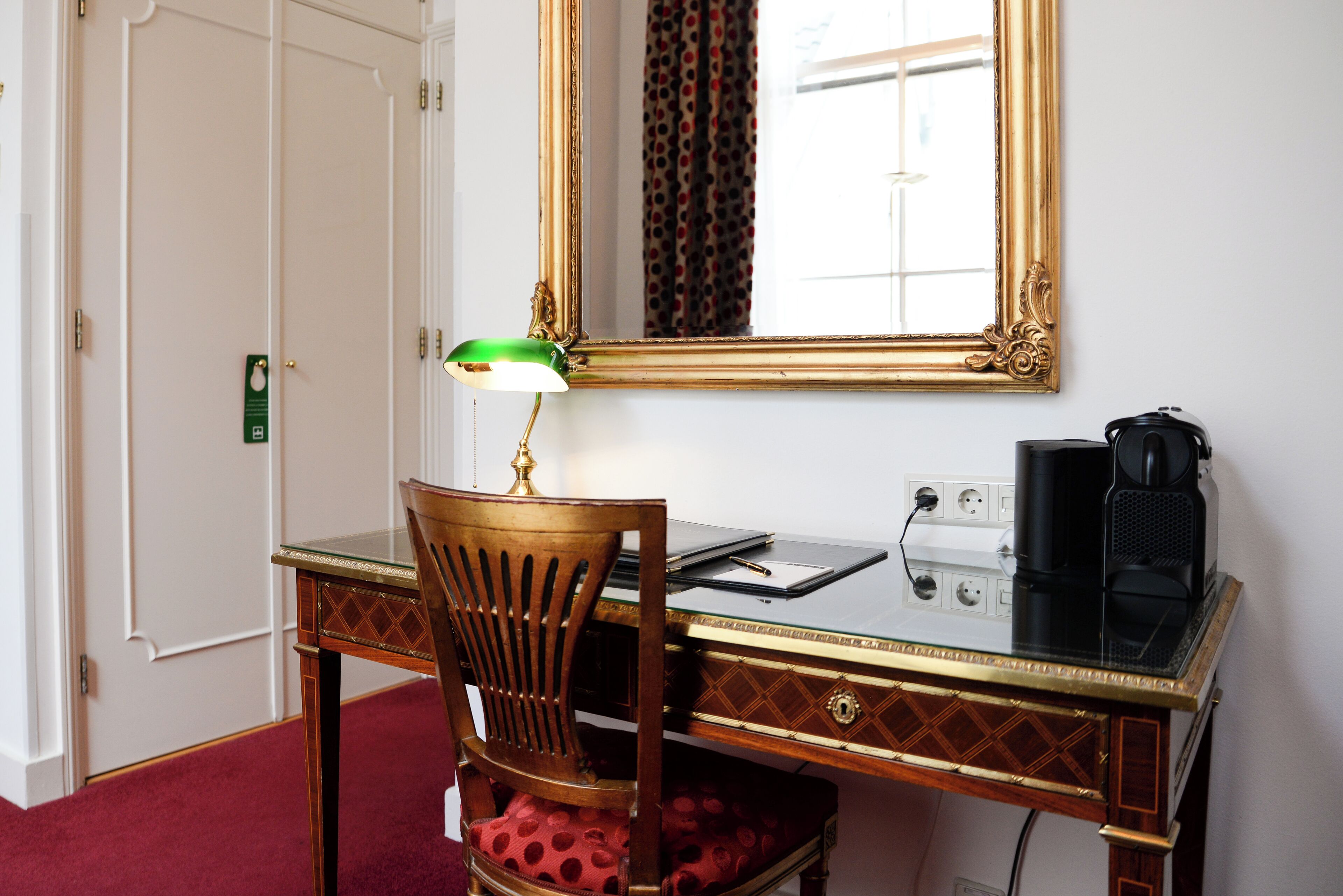 Quarto de hotel com escrivaninha de madeira, luminária de mesa verde, cafeteira preta e espelho com moldura decorada.