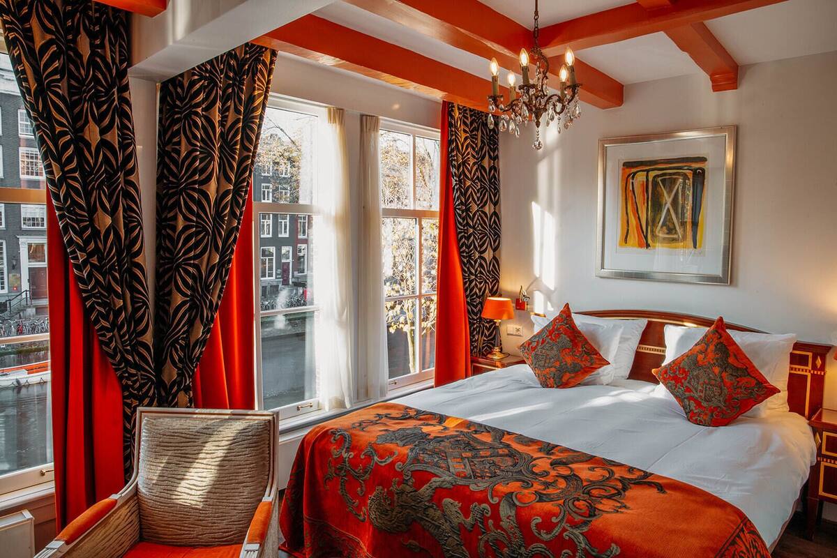 Quarto de hotel com uma cama grande, cortinas com padrão em vermelho e preto, um lustre e vista para a cidade.