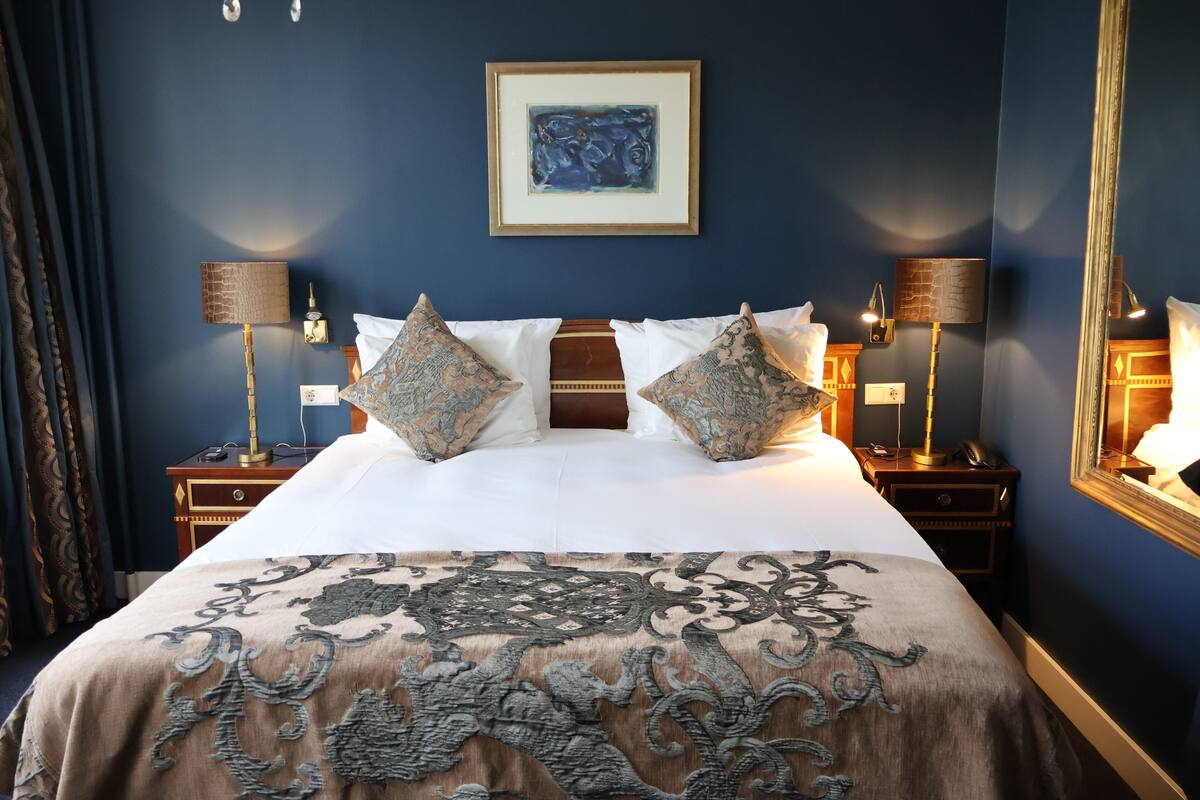 Cama bem arrumada com almofadas estampadas, uma grande obra de arte emoldurada acima e duas luminárias de cabeceira em mesas de cabeceira de madeira.
