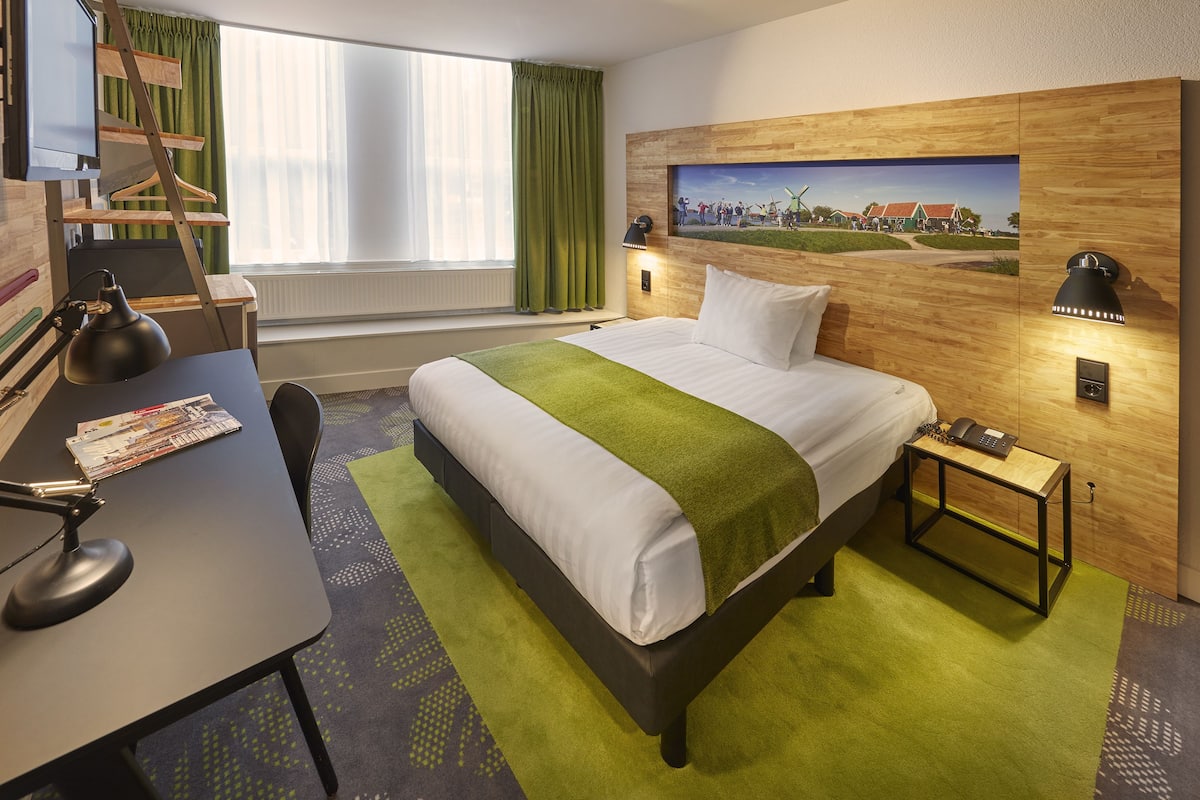 A hotel room with a bed, desk, chair, lamp, and a painting on the wall.