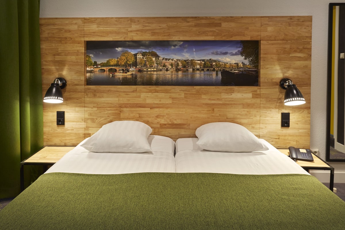 A hotel room with a wooden headboard, a large cityscape painting, two bedside lamps, and a green bedspread.
