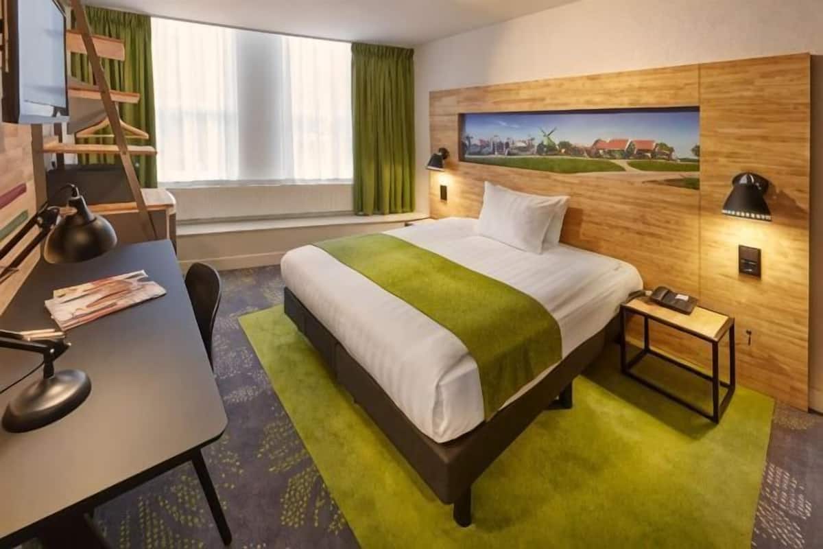 Quarto de hotel com cama, escrivaninha, cadeira, luminária e um quadro na parede.