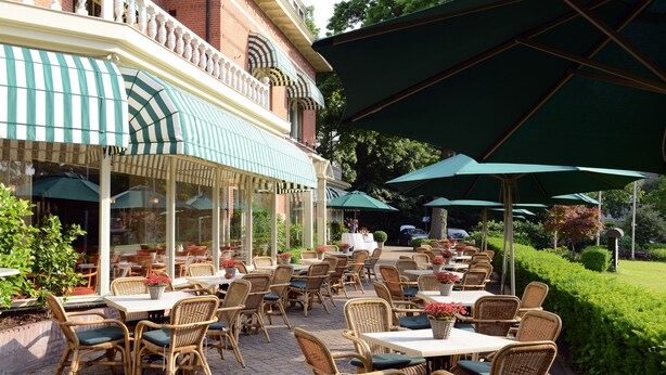 Een terras buiten met rieten stoelen en tafels, groene parasols, en een gebouw met bakstenen muren en een gestreepte luifel.