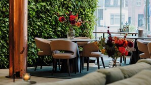 An indoor seating area with tables and chairs, a green wall with plants, and a window with a view of buildings. - Gooiland Hotel (Hilversum)