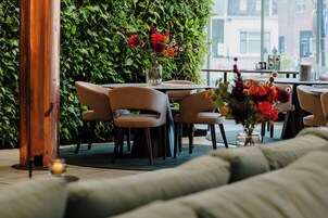 Un área de estar interior con mesas y sillas, una pared verde con plantas y una ventana con vistas a edificios.