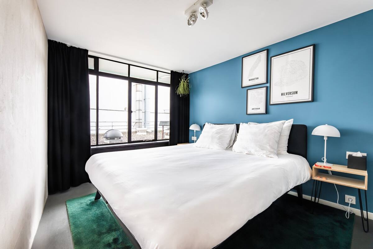 A modern hotel room with a large bed, a nightstand with a lamp, and a window with black curtains.