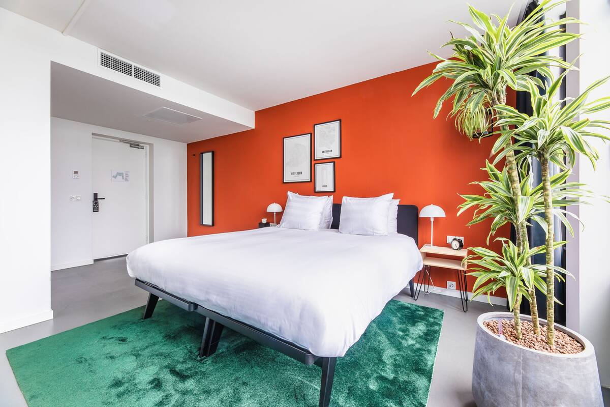 A modern hotel room with a large bed, a green rug, a potted plant, and a bright orange accent wall.
