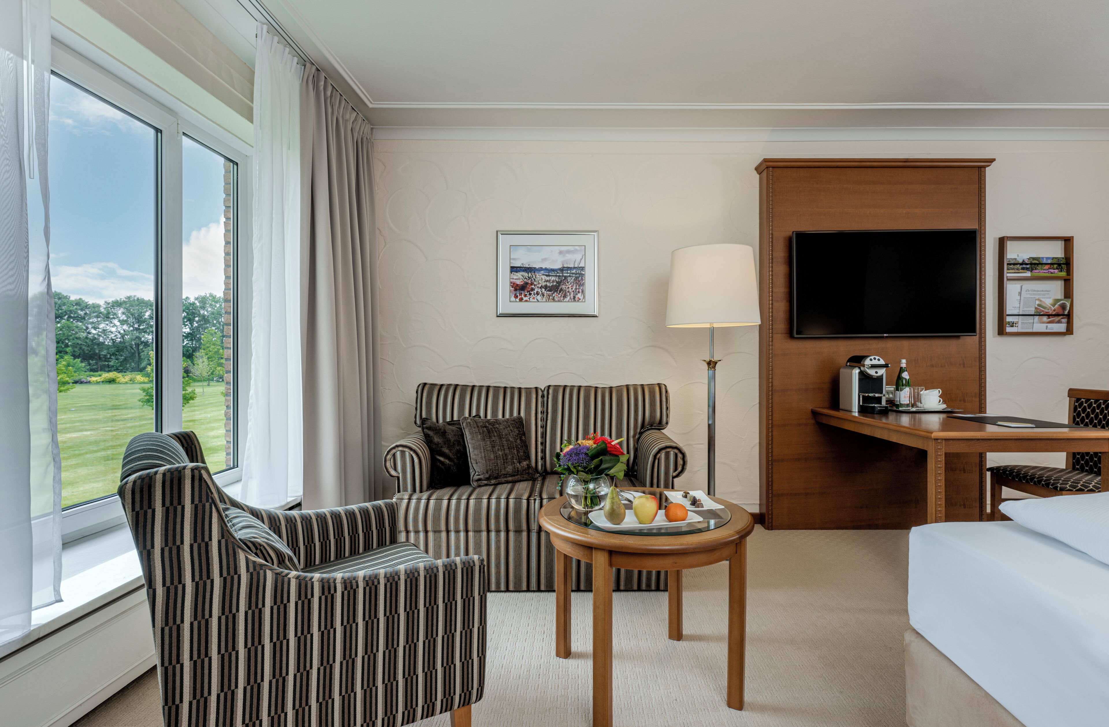 Habitación de hotel con sofá, sillón, mesa de comedor con fruta, televisor y escritorio con lámpara.