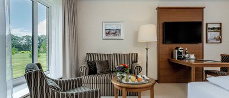A hotel room with a sofa, armchair, dining table with fruit, a TV, and a desk with a lamp.