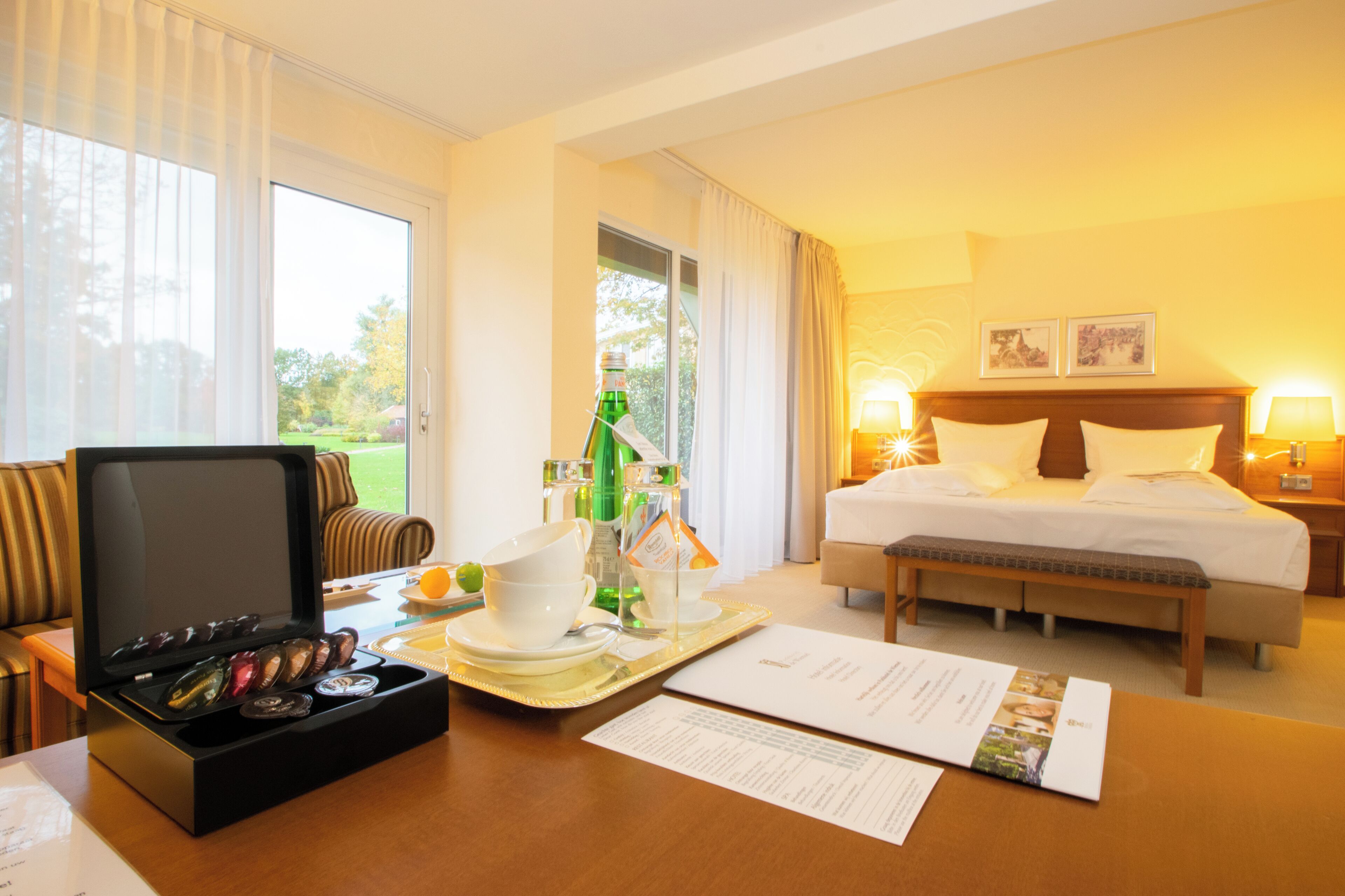 A hotel room with a bed, bedside lamps, a desk with a tray of breakfast items, a television, and a window with curtains.
