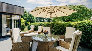 An outdoor seating area with wicker chairs and a glass table, surrounded by lush greenery and a modern building. - Fletcher Parkhotel De Wiemsel (Ootmarsum)