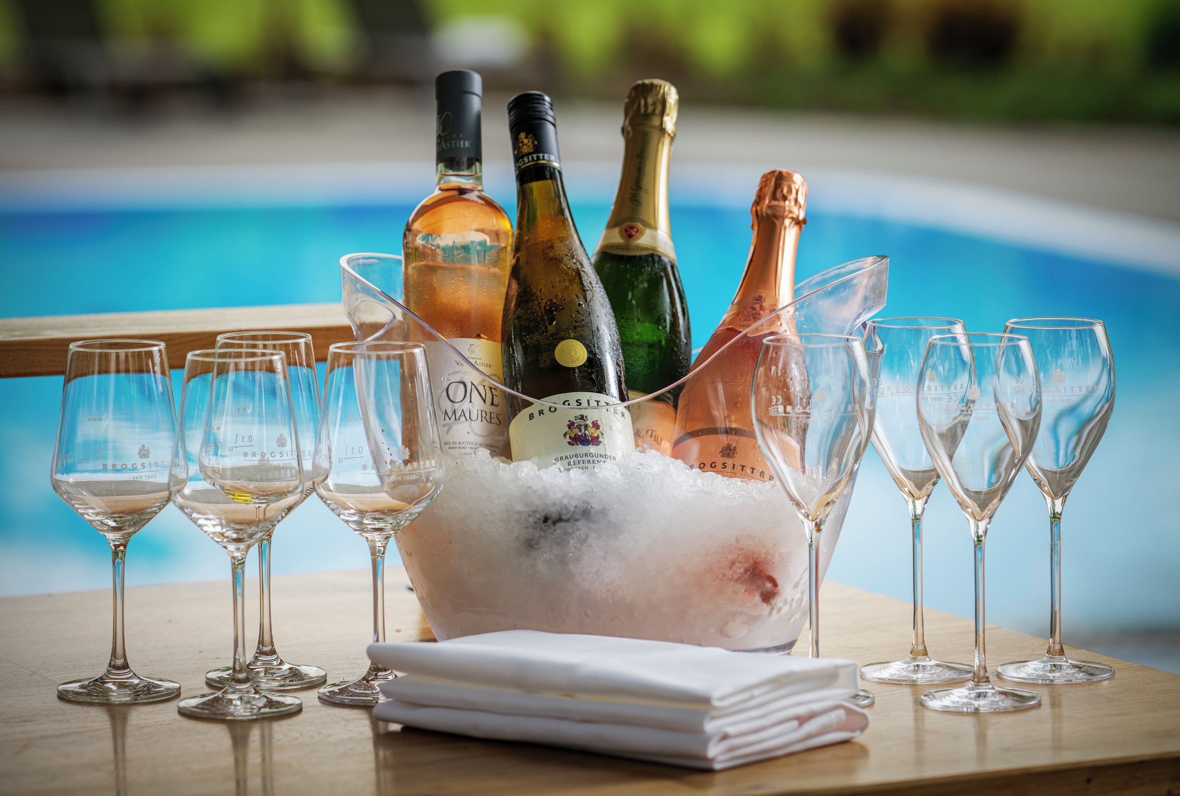 A poolside setting with a champagne bucket filled with ice and several wine glasses, some with bottles of champagne.