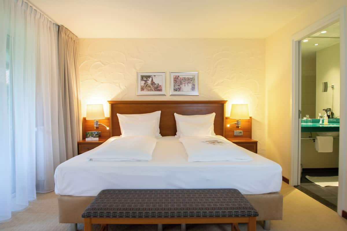 Une chambre d’hôtel avec un grand lit, deux tables de chevet avec des lampes, un banc et deux tableaux encadrés au mur.
