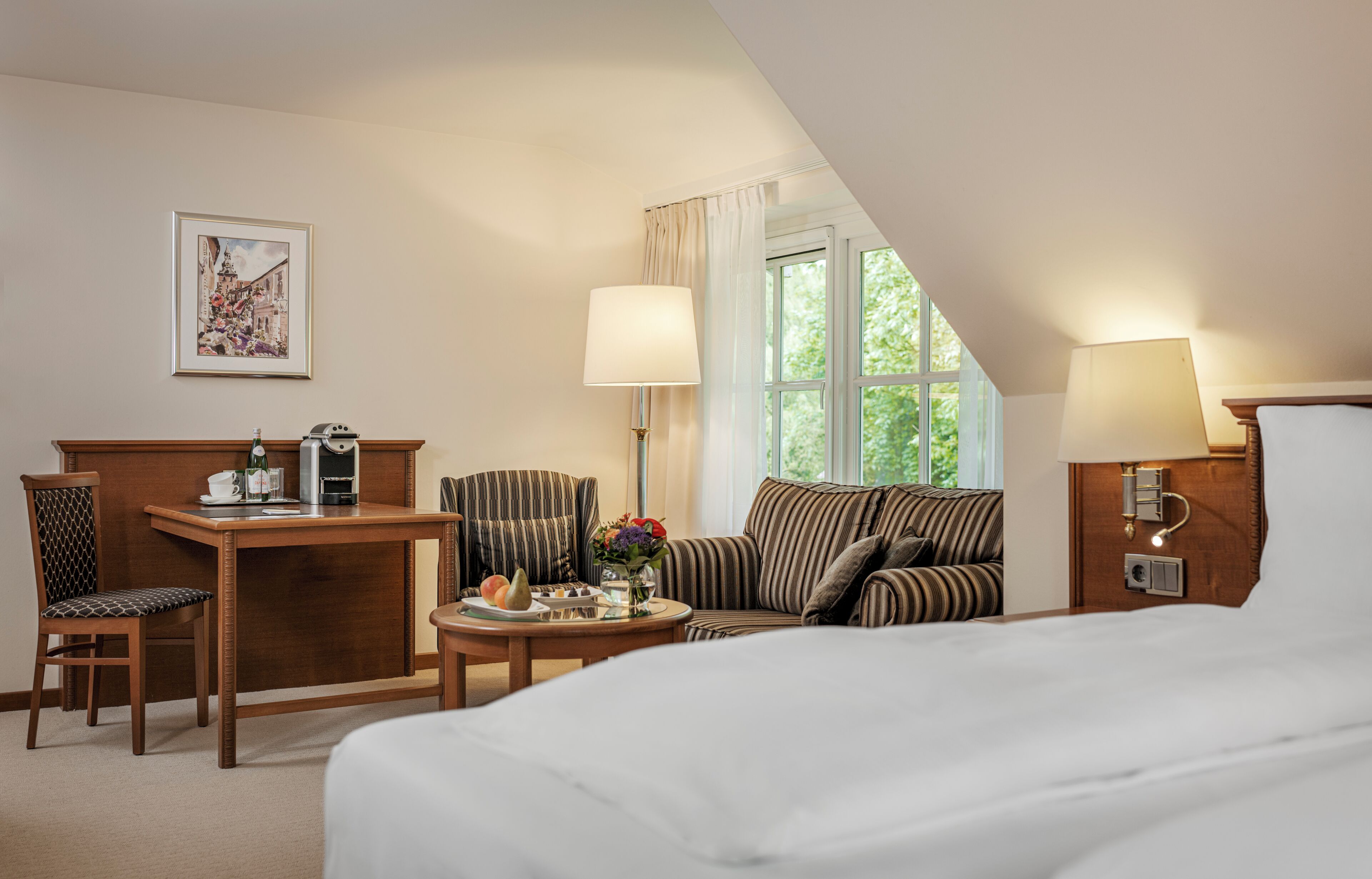 Habitación de hotel con cama, escritorio con cafetera, sillón a rayas, sofá y ventana con cortinas.