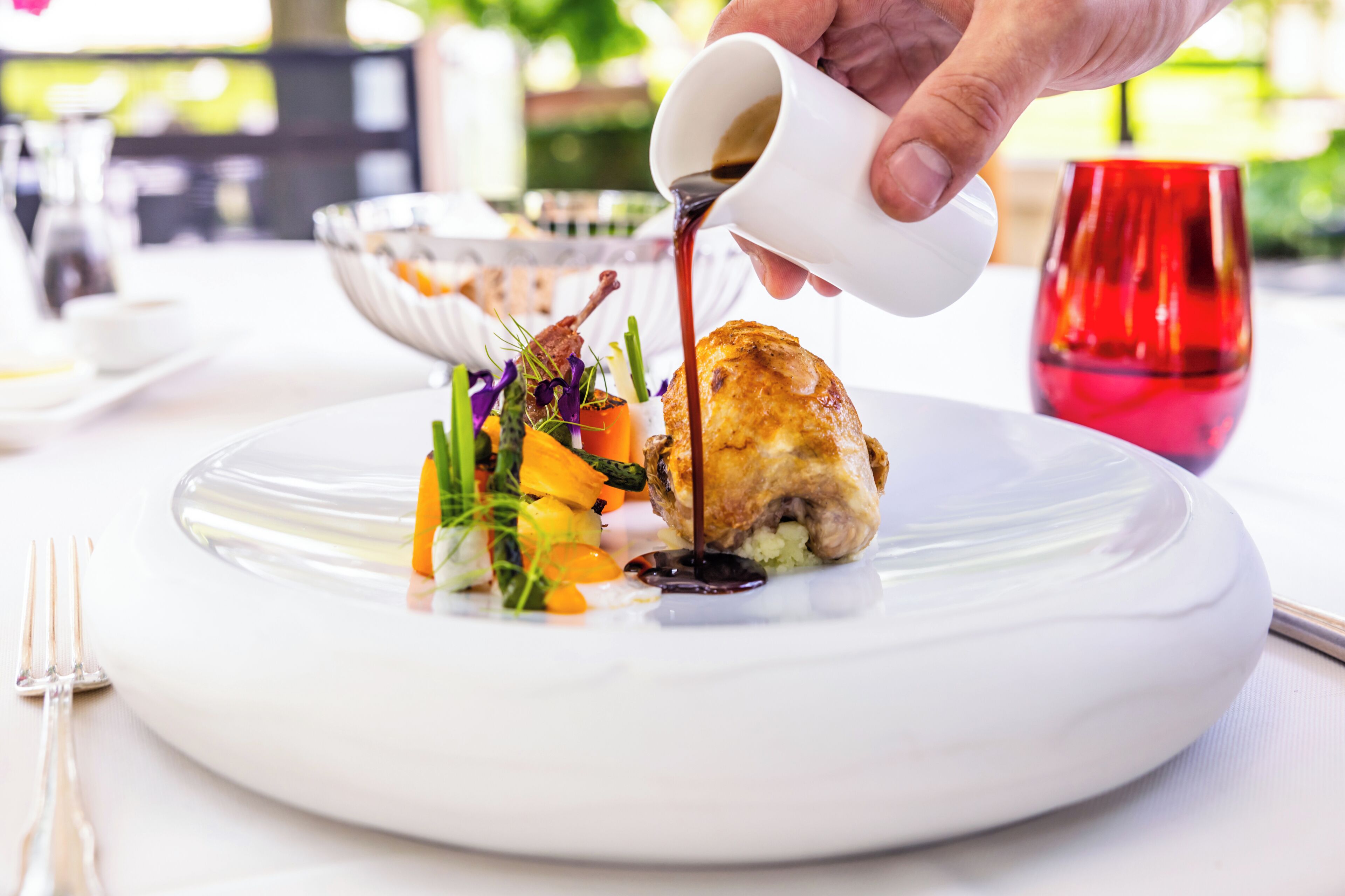 A white plate with a roasted chicken leg, vegetables, and sauce being poured from a white cup.