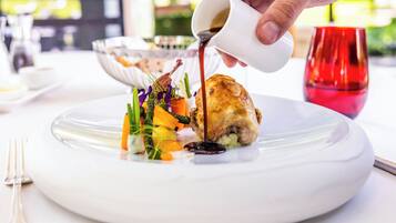 A white plate with a roasted chicken leg, vegetables, and sauce being poured from a white cup.